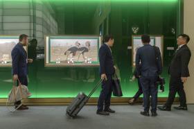 Parete verde retroilluminata con vetrine nelle quali sono esposti orologi da polso; davanti, uomini con valige trolley