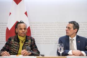 Una donna in blazer e dolcevita casual e un uomo in abito formale seduti a un tavolo di conferenza stampa; bandiera CH dietro