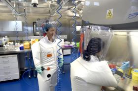 Interno di laboratorio con due laboratoriste completamente coperte da indumenti di protezione e casco ventilato di protezione