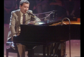 Un uomo in abito e cravatta seduto al pianoforte ma di lato, con una sola mano sulla tastiera; canta