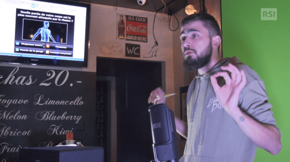 Giovane uomo parla davanti a un microfono verso una telecamera all interno di un piccolo bar; sul fondo schermo con domande quiz