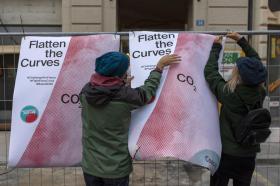 Due giovani donne appendono un manifesto con scritto Flatten the curves a una recinzione.