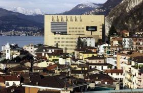 Casino di Campione visto di lato in giornata soleggiata, con abitato attorno e lago e Alpi sullo sfondo