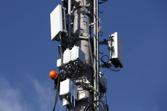 Immagine di un antenna di telefonia mobile con 5G e 4G; operaio con casco arancione effettua riparazione.
