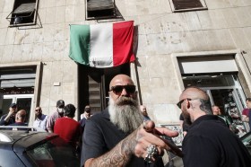 Uomo con testa rasata, barba lunga e occhiali da sole punta il dito contro la camera: dietro, palazzo con bandiera italiana