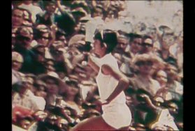 Un atleta in tenuta bianca corre in salita reggendo una fiaccola; dietro, pubblico da stadio