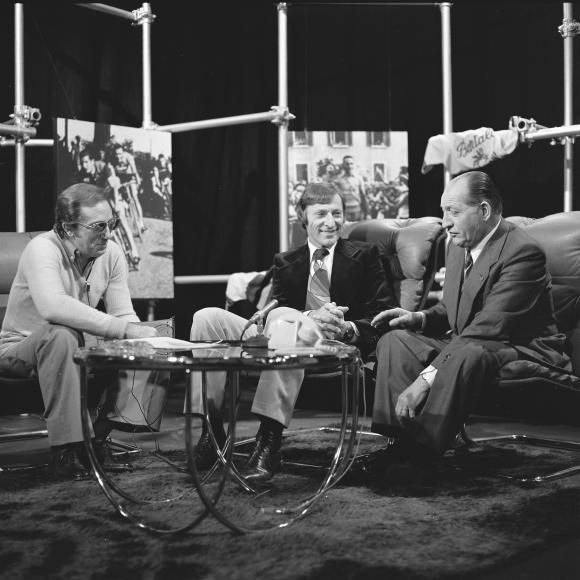 b/n Tre uomini seduti su due divanetti attorno a un tavolino in una scenografia televisiva; uno parla altri ascoltano