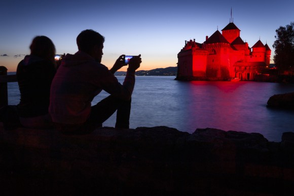 Castello in riva a un lago completamente illuminato di rosso; persone fotografano col cellulare