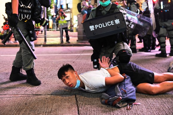 Giovane a terra in strada di città; poliziotto munito di scudo lo tiene a terra con ginocchio