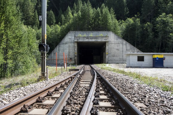 Portale in cemento armato di tunnel ferroviario visto dai binari antistanti; bosco attorno