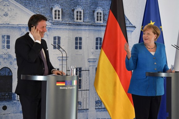 Conte sorride mentre ascolta una traduzione, Merkel lo guarda facendogli un cenno; immagine di un castello e bandiere sul fondo