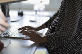 Di profilo, donna incinta (indossa camicia scura a pois) lavora al computer a scrivania d ufficio
