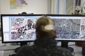 Donna (di schiena, sfocata) lavora a un computer con doppio schermo; su uno foto aerea, sull altro mappa