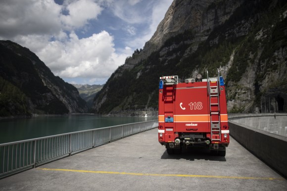 Camion dei pompieri parcheggiato su ponte accanto a lago artificiale in ambiente alpino.