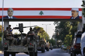 Passerella pedonale coperta da lunga bandiera libanese con volto di un uomo; convoglio militare transita al di sotto