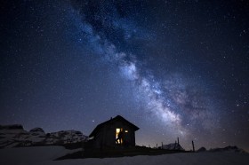 Cielo stellato visto in condizioni di luce ideale in ambiente alpino; Piccola baita con finestra illuminata in primo piano