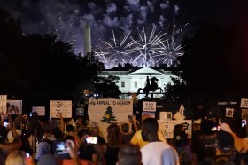 Folla di manifestanti con cartelli vari in penombra in primo piano; Casa Bianca e fuochi artificiali a fuoco sul fondo