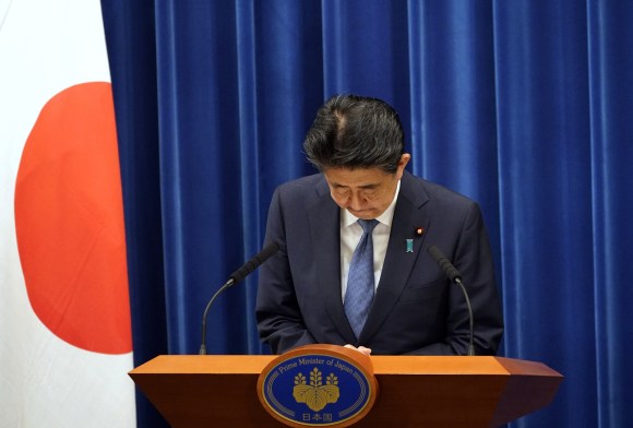 Shinzo Abe al pulpito guarda i suoi appunti a testa bassa con espressione di rammarico; bandiera giapponese accanto