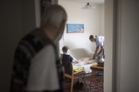 un anziano osserva un infermiera che si occupa di sua moglie in salotto.