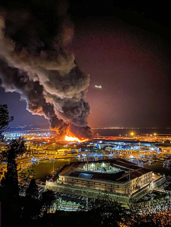 Zona industriale portuale vista di notte con visibile rogo al centro e colonna di fumo denso che si alza