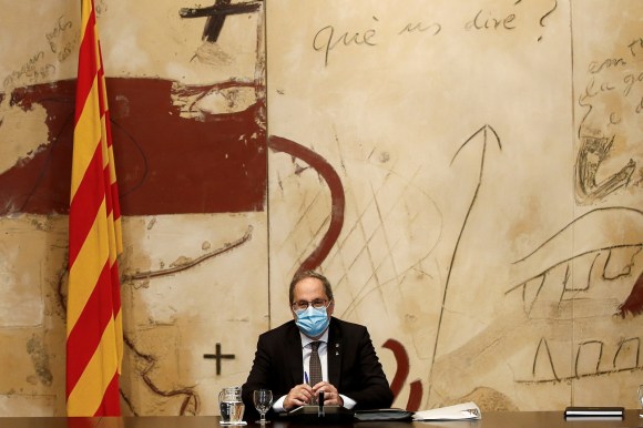 Uomo in abiti formali, con mascherina, seduto a una scrivania sovrastata da grande murale artistico e bandiera catalana