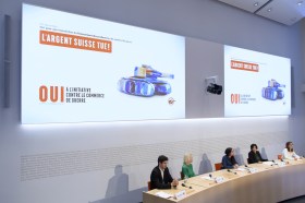 Quattro donne e un uomo a un tavolo di conferenza stampa; alle loro spalle effige di carro armato composto con banconote