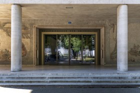 Entrata di un edificio con colonne e mosaici decorativi, porte scorrevoli nelle quali si specchia un parco