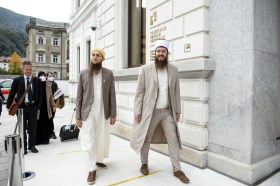 Due uomini con barbe lunghe e copricapo, uno con tunica, camminano davanti a edificio con scritta Tribunale Penale federale