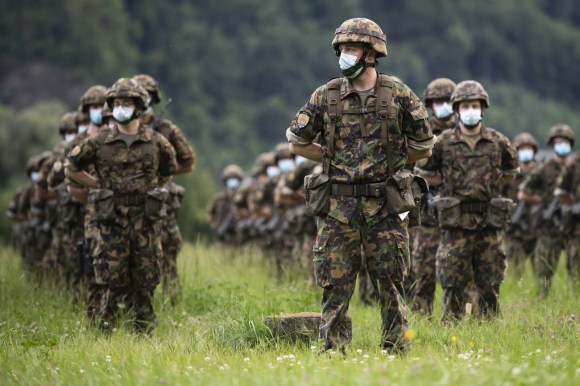Decine di militari in formazione e posizione di riposo su un prato; tutti indossano la mascherina
