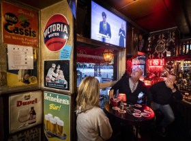 Una donna e due uomini al tavolo di un pub guardano due politici che annunciano qualcosa in tv