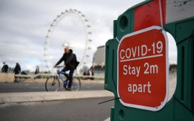 Primo piano di transenna con cartello Covid-19 Stay 2m apart ; si intravvedono un ciclista e la London Eye