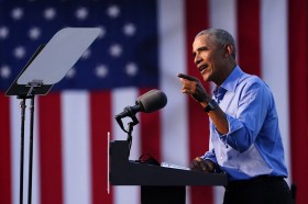 Barack Obama su un palco, a un pulpito con due microfoni, ripreso di lato; sullo sfondo bandiera USA