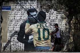 Muro cittadino ombreggiato da pianta con dipinto che raffigura Pelé e Alì che s abbracciano; sul fondo scritte Get up, Stand up