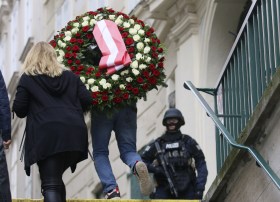 due persone portano una corona mortuaria sotto lo sguardo di un poliziotto