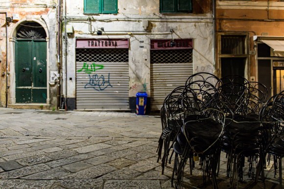 Piazzetta di centro storico con serrande abbassate e sedie impilate; scritta Trattoria