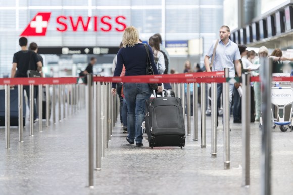 Interno di aeroporto, con nastri per guidare le code al check-in; persone con bagagli in lontananza, scritta Swiss