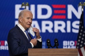 Biden si toglie la mascherina mentre si avvicina a un pulpito; logo Biden-Harris sul fondo, bandiera USA accanto