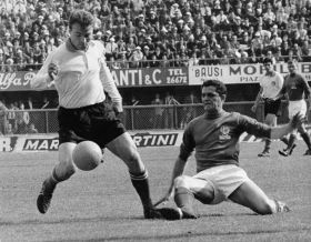 Un giocatore di calcio ne scarta un altro in una vecchia foto in bianco e nero. Sul fondo, spalti, pubblicità e pubblico