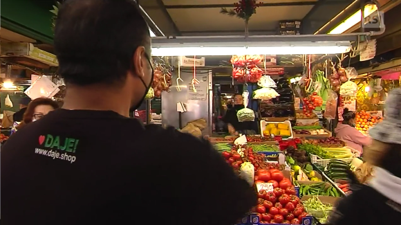 Banco di mercato con frutta e verdura. Di spalle, un giovane con maglietta con scritto www.daje.shop