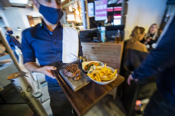 Un cameriere con mascherina porta tre pietanze su un vassoio in un ristorante affollato