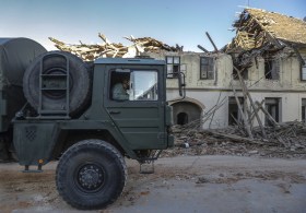 Camion militare transita davanti a fila di case contigue parzialmente distrutte