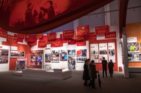 Sala espositiva con foto, teche e numerose bandiere rosse con simboli comunisti; visitatori con mascherina