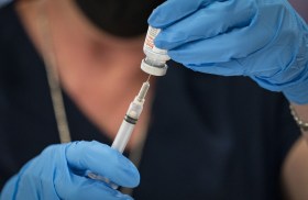Primo piano di due mani con guanti azzurri che estraggono da una fiala, con una siringa, una dose di vaccino