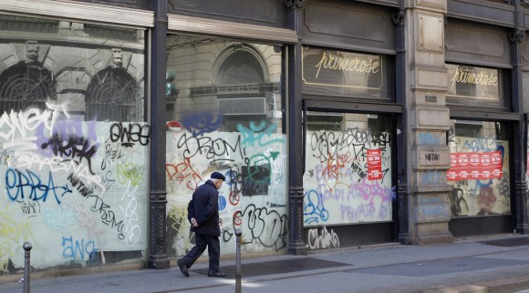 Un anziano signore cammina in centro di Milano davanti a negozi chiusi per fallimento.
