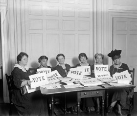 donne sedute a un tavolo con cartelloni con la scritta voto