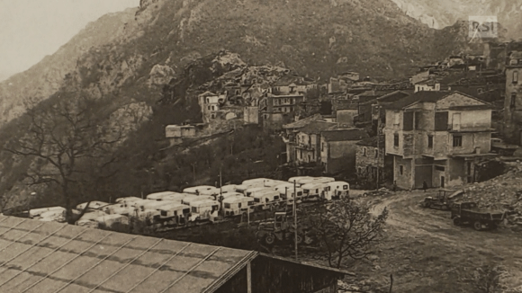 Immagine di un villaggio ai piedi di una montagna con, ai margini, una fila di roulotte parcheggiate.
