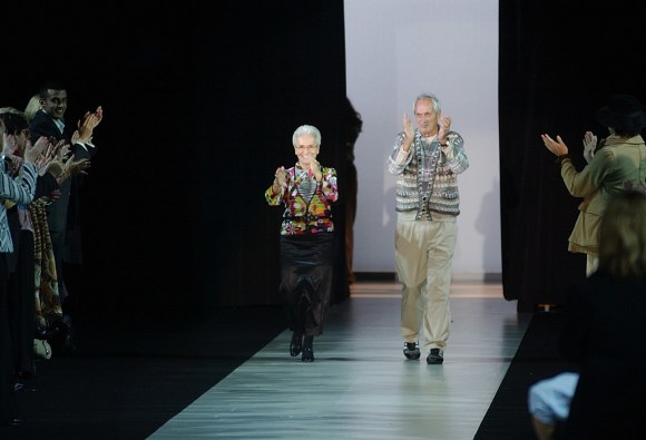 Una donna e un uomo sulla settantina-ottantina camminano su una passerella; indossano abiti di maglieria colorati