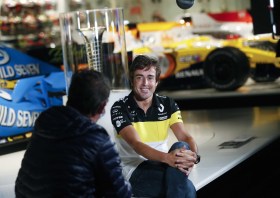 Fernando Alonso, seduto sul bordo di un palco sul quale sono esposte due monoposto, parla con un giornalista