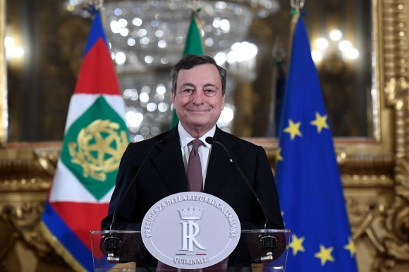 Draghi in piedi al pulpito col simbolo della Repubblica italiana, dietro: bandiere italiana, UE e della Presidenza della Rep