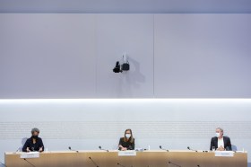 Due donne e un uomo, molto distanti le une dall altro, a un tavolo di sala stampa; fondo completamente bianco.